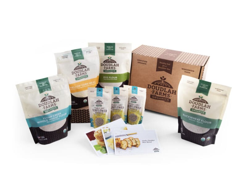 A Coolbeans Basket from Doudlah Farms showcases an array of organic products, including assorted flours in bags, seeds in packets, and a cardboard box, all elegantly arranged on a white background. The addition of recipe cards provides an inspiring touch to the presentation.