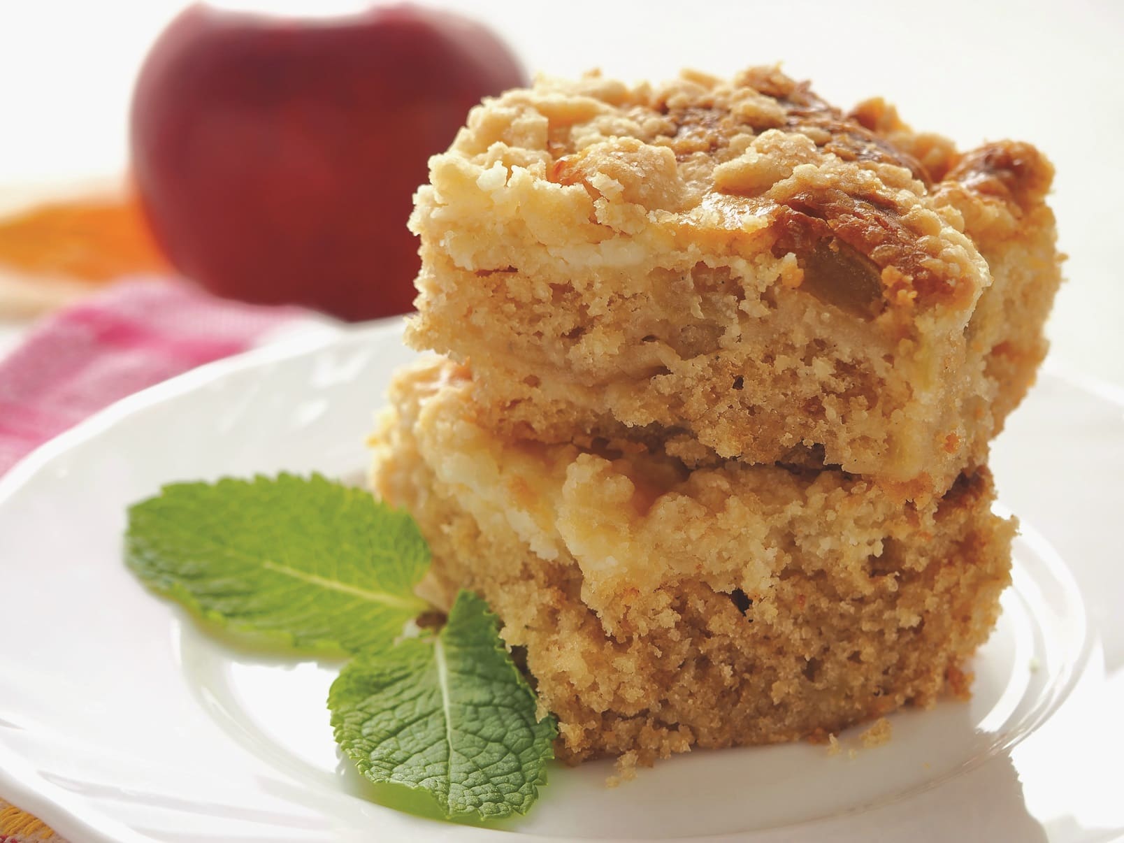 Two square pieces of apple crumb cake, with a hint of cheddar, are stacked on a white plate and garnished with fresh mint leaves. A red apple is blurred in the background, adding a touch of color.