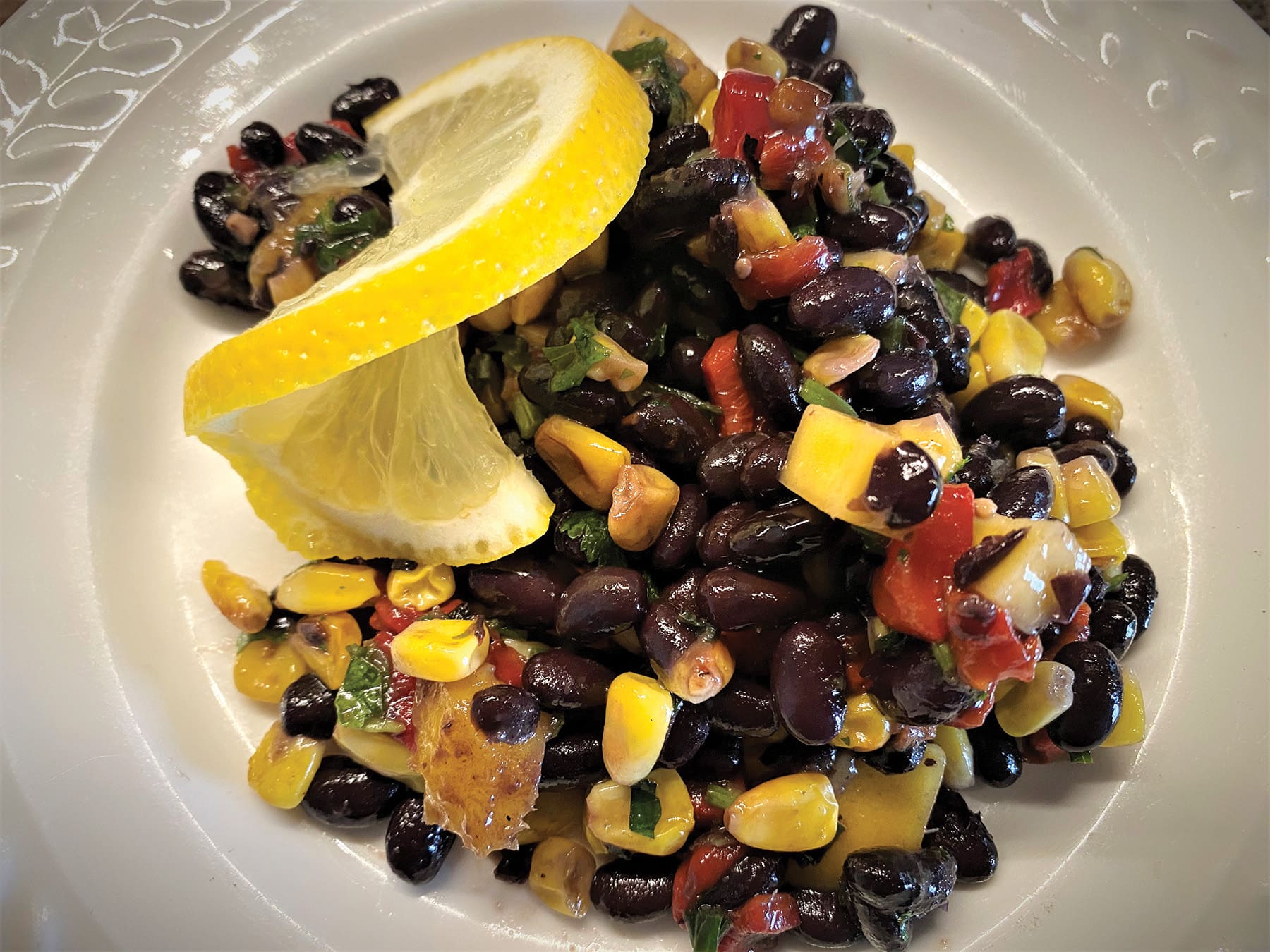 A vibrant salad featuring black beans, corn, diced red and yellow peppers, garnished with fresh cilantro and lemon slices, served with a side of garlic cheddar beer bread on a white plate.