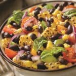 A colorful salad in a bowl features black beans, grilled corn, cherry tomatoes, red onion slices, avocado, and fresh basil leaves enhanced with a hint of garlic. The ingredients are mixed together, creating a vibrant and appetizing dish.