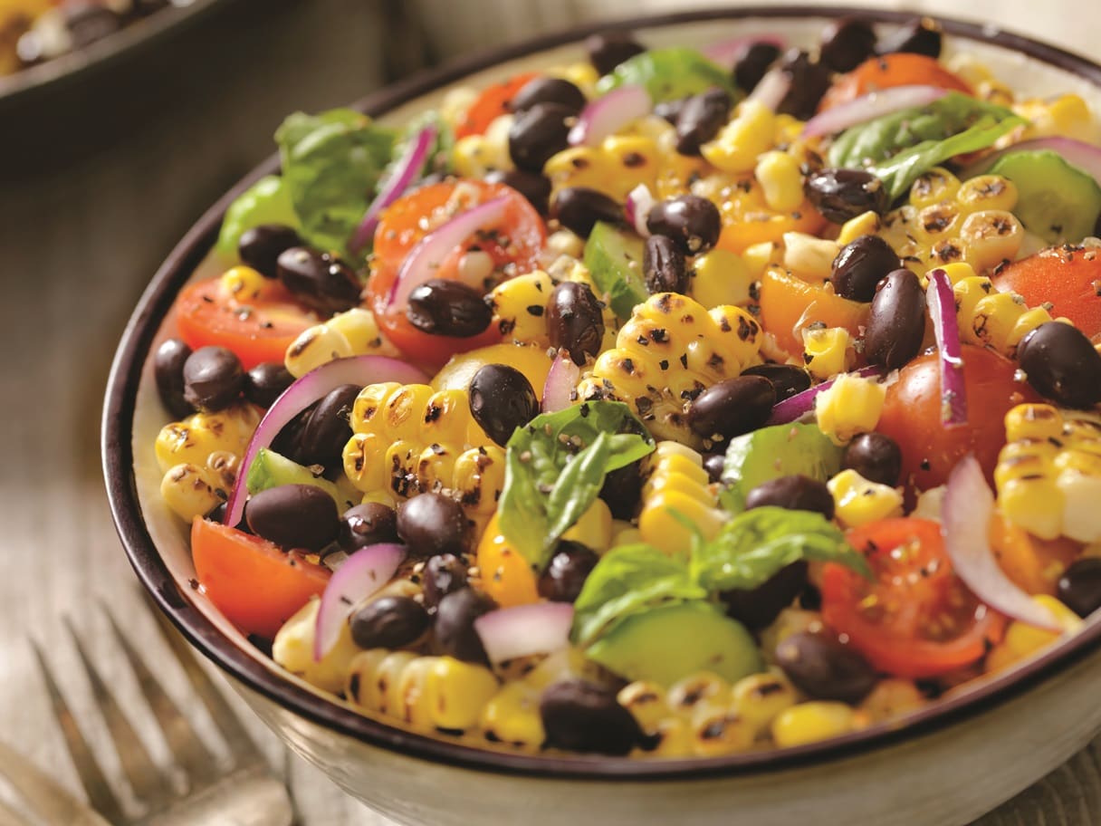 A colorful salad in a bowl features black beans, grilled corn, cherry tomatoes, red onion slices, avocado, and fresh basil leaves enhanced with a hint of garlic. The ingredients are mixed together, creating a vibrant and appetizing dish.