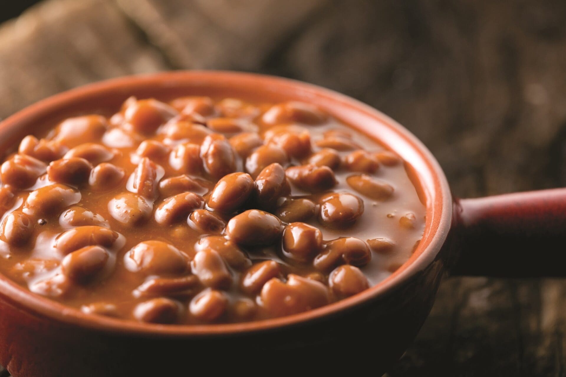 Close-up of a brown ceramic bowl filled with baked beans in a rich, thick sauce, garnished with a hint of garlic. The bowl is set on a wooden surface, giving a rustic feel to the image. The glossy beans look appetizing next to slices of savory beer bread.