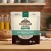 A package of Doudlah Farms Organics Buckwheat Flour, certified organic and gluten-free from Wisconsin, is prominently displayed on a kitchen counter, with a loaf of bread and glasses of juice enhancing the rustic kitchen scene.