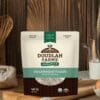 A Douglah Farms Organics Buckwheat Flour bag rests on a wooden table, featuring white packaging with green and brown accents. Labels confirm its certified organic and gluten-free, while blurred kitchen items in the background suggest culinary possibilities.