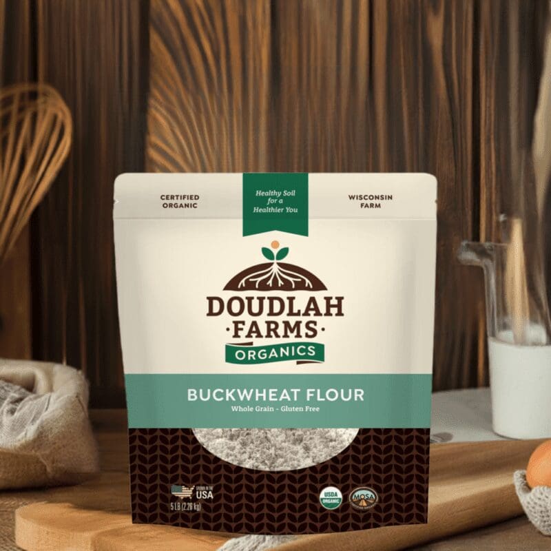 A Douglah Farms Organics Buckwheat Flour bag rests on a wooden table, featuring white packaging with green and brown accents. Labels confirm its certified organic and gluten-free, while blurred kitchen items in the background suggest culinary possibilities.