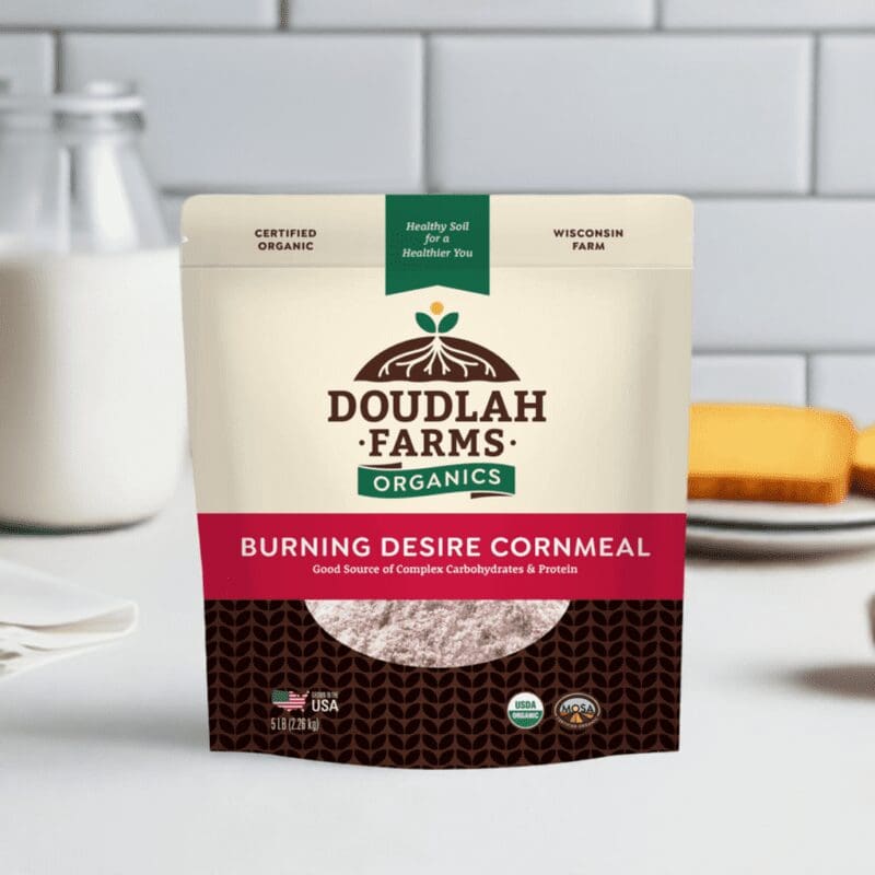 The Burning Desire Cornmeal from Doudlah Farms Organics, made in Wisconsin, takes center stage on the kitchen counter. A tempting slice of cake and a glass of milk behind it promise a deliciously fiery touch to your cooking adventures.