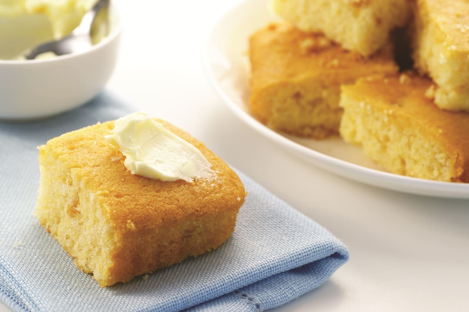 A square piece of cornbread topped with butter sits on a blue napkin, accompanied by hints of cheddar. In the background, more pieces of cornbread rest on a white plate. To the left, a bowl with a spoon is partially visible.