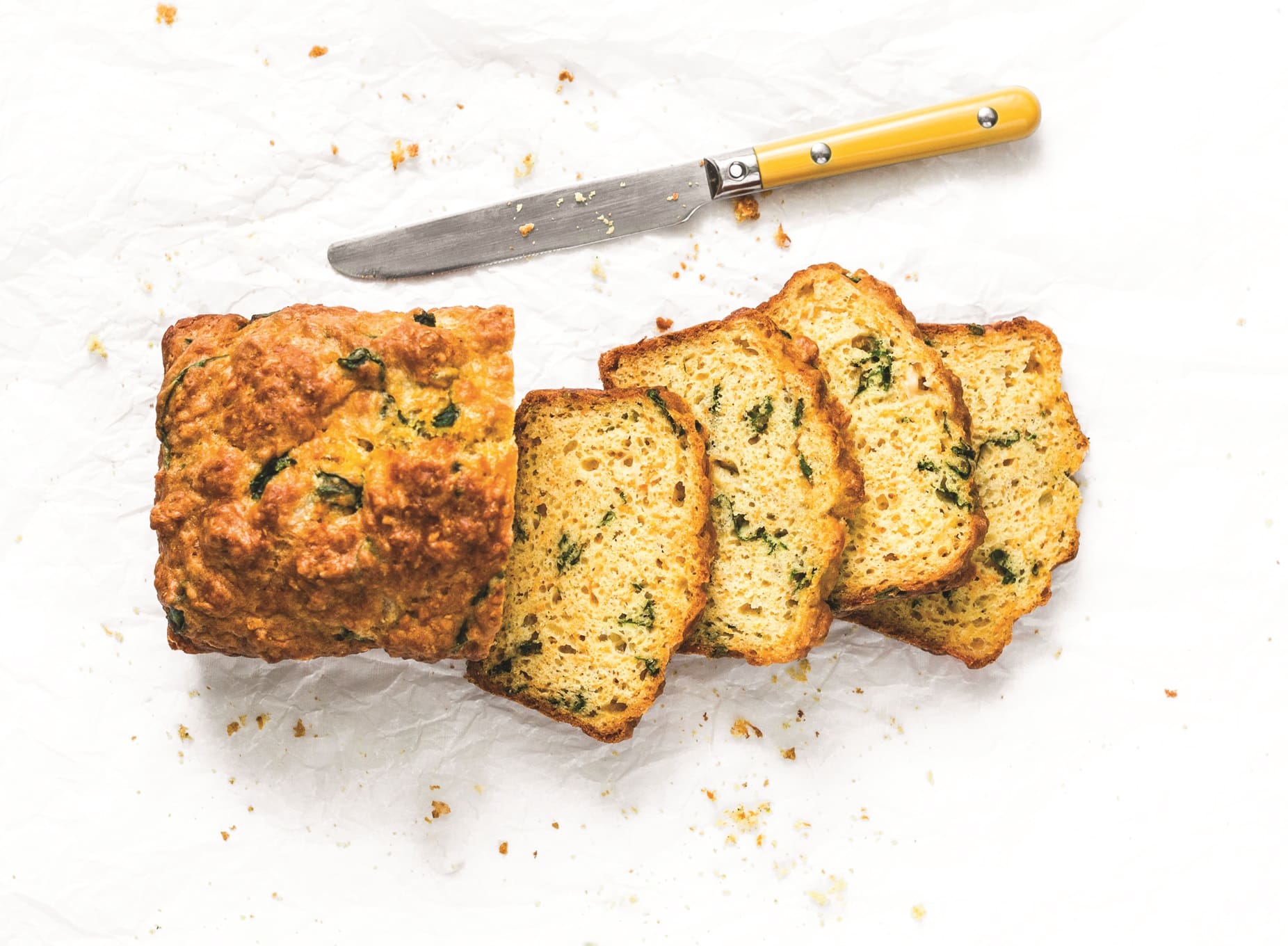 A loaf of herb-infused beer bread partially sliced, with a knife featuring a yellow handle resting beside it. The bread is placed on crinkled white parchment paper, with scattered breadcrumbs nearby.