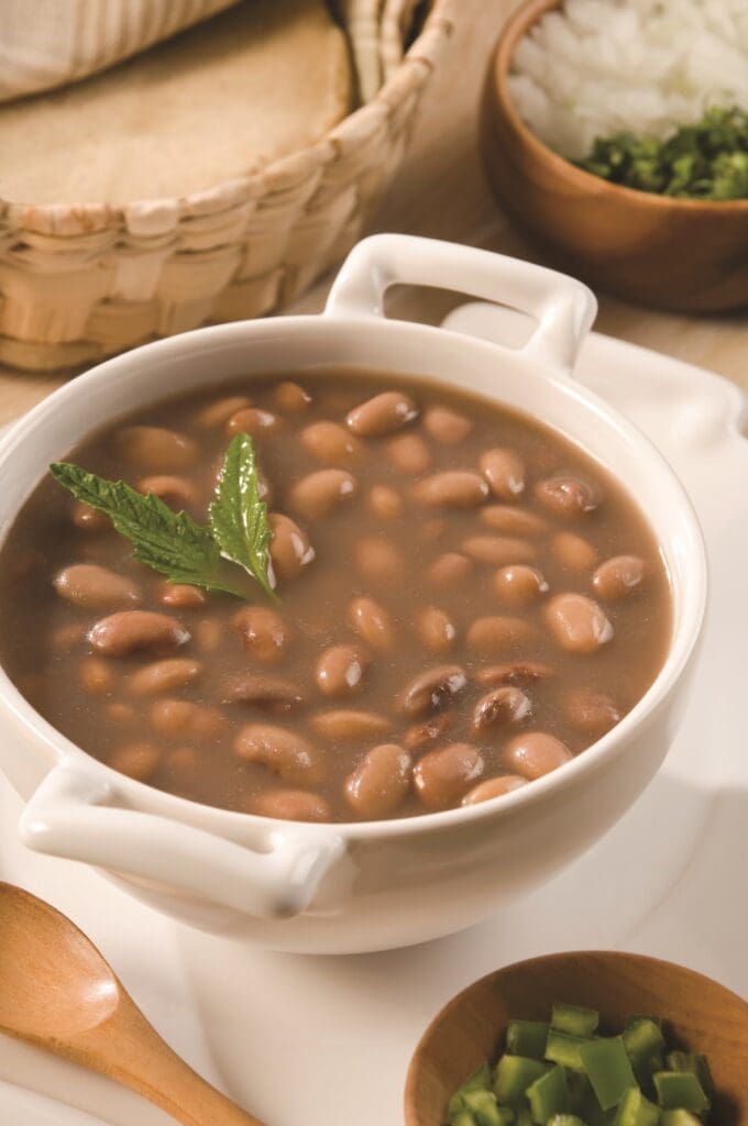 A white bowl filled with savory bean soup, garnished with a fresh mint leaf, rests on a plate. Encircling the bowl are wooden containers holding diced onions and green peppers, alongside a spoon and a basket of bread or tortillas. Perfectly crafted in an Instant Pot for that quick yet rich flavor.