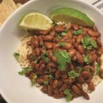 A bowl of seasoned brown rice topped with pinto beans, garnished with fresh cilantro. Two lime wedges rest on the side. Tortilla chips are partially visible in the background on a striped surface, hinting at a recipe that's as easy as using your Instant Pot.