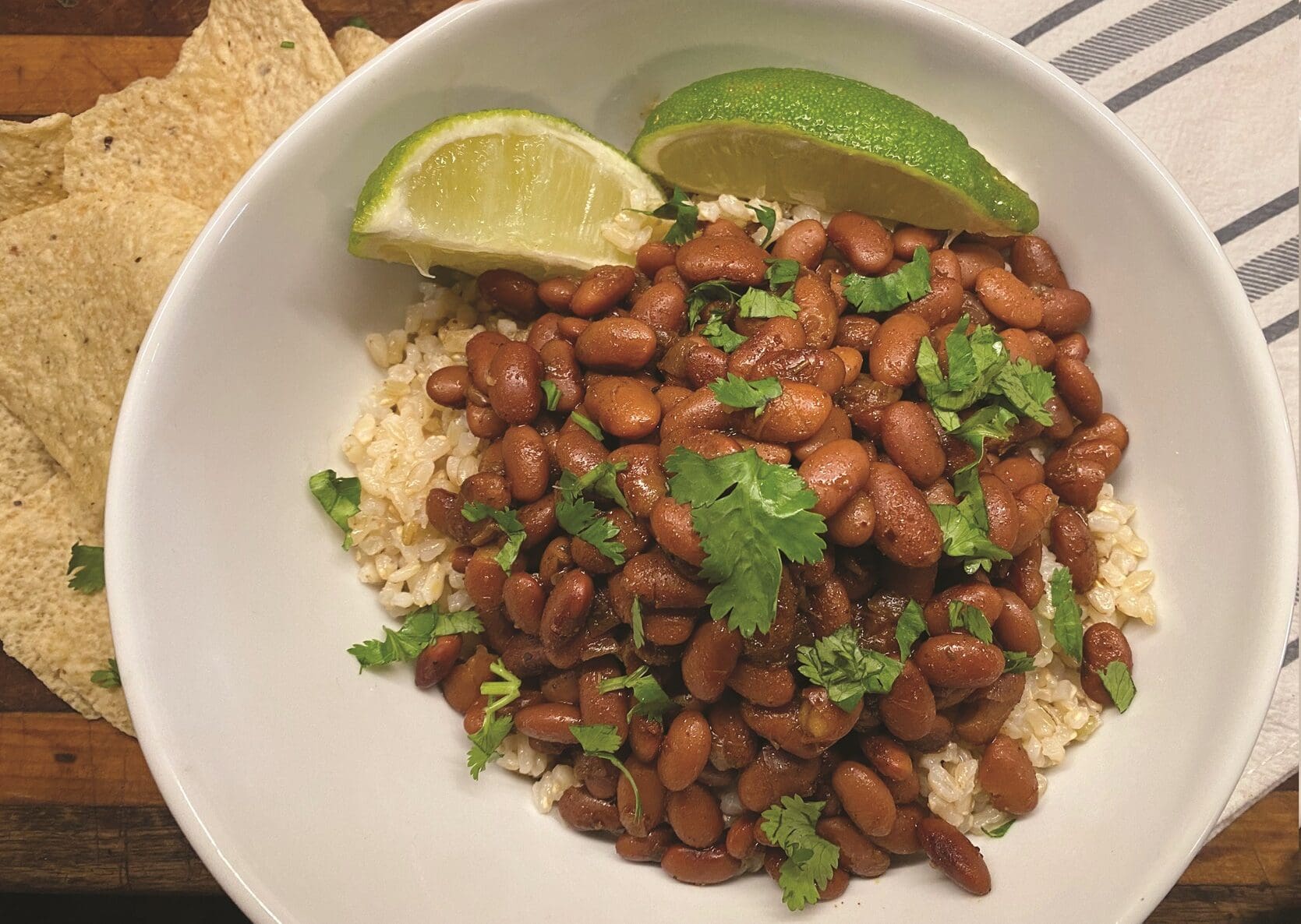 A bowl of seasoned brown rice topped with pinto beans, garnished with fresh cilantro. Two lime wedges rest on the side. Tortilla chips are partially visible in the background on a striped surface, hinting at a recipe that's as easy as using your Instant Pot.