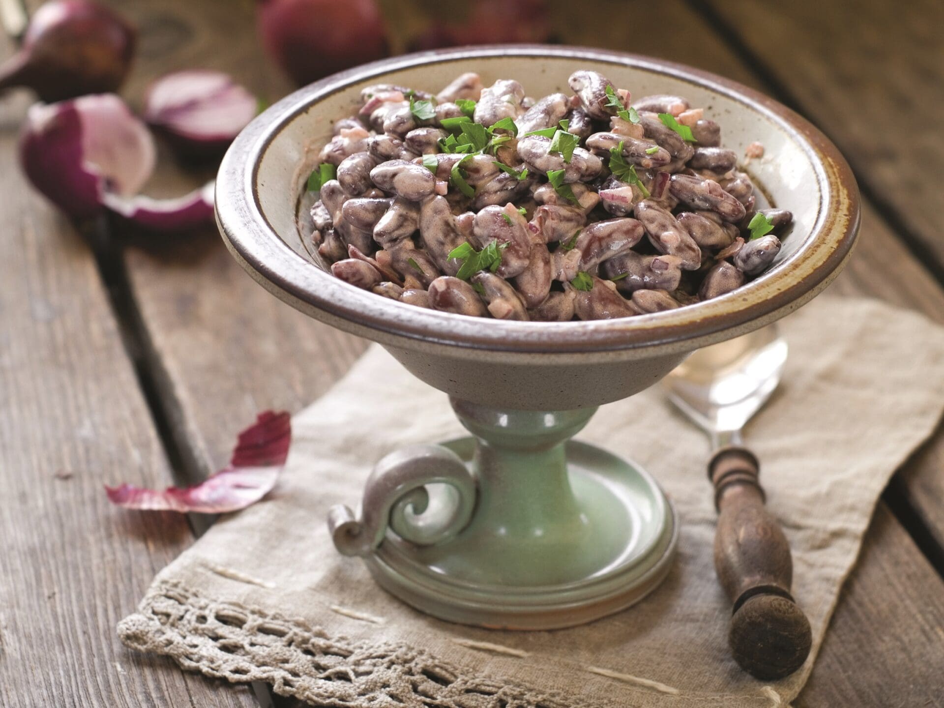 A vintage pedestal bowl brimming with a creamy kidney bean salad, garnished with fresh herbs. The bowl rests on a rustic wooden table draped with a linen cloth, accompanied by a nearby fork. In the background, vibrant red onion halves add a splash of color.