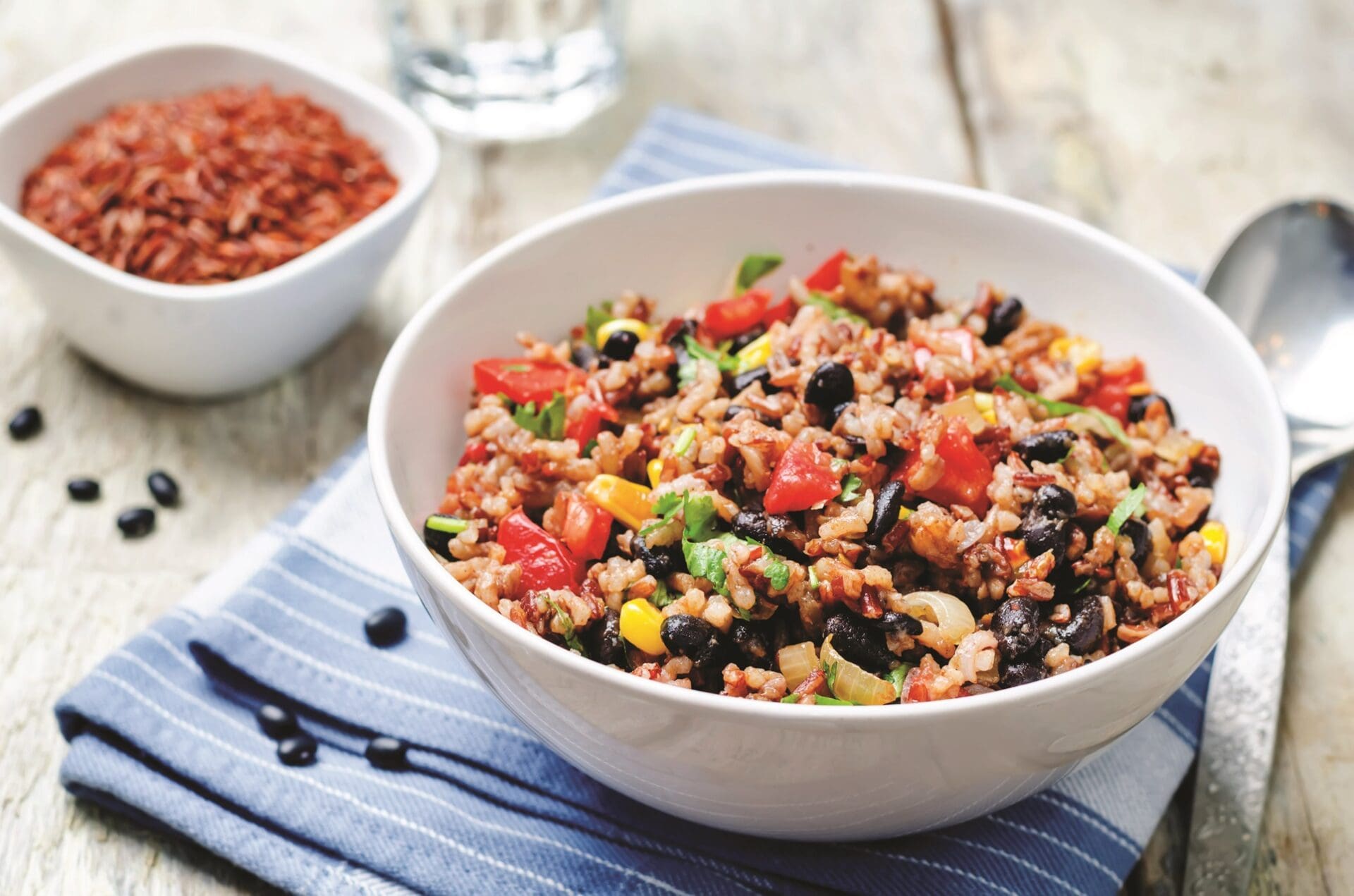 A bowl of mixed rice salad with Latin black beans, corn, tomatoes, and fresh herbs sits on a blue striped cloth. Embracing the essence of Latin cuisine, a small bowl of red rice is in the background next to a spoon on a wooden surface.