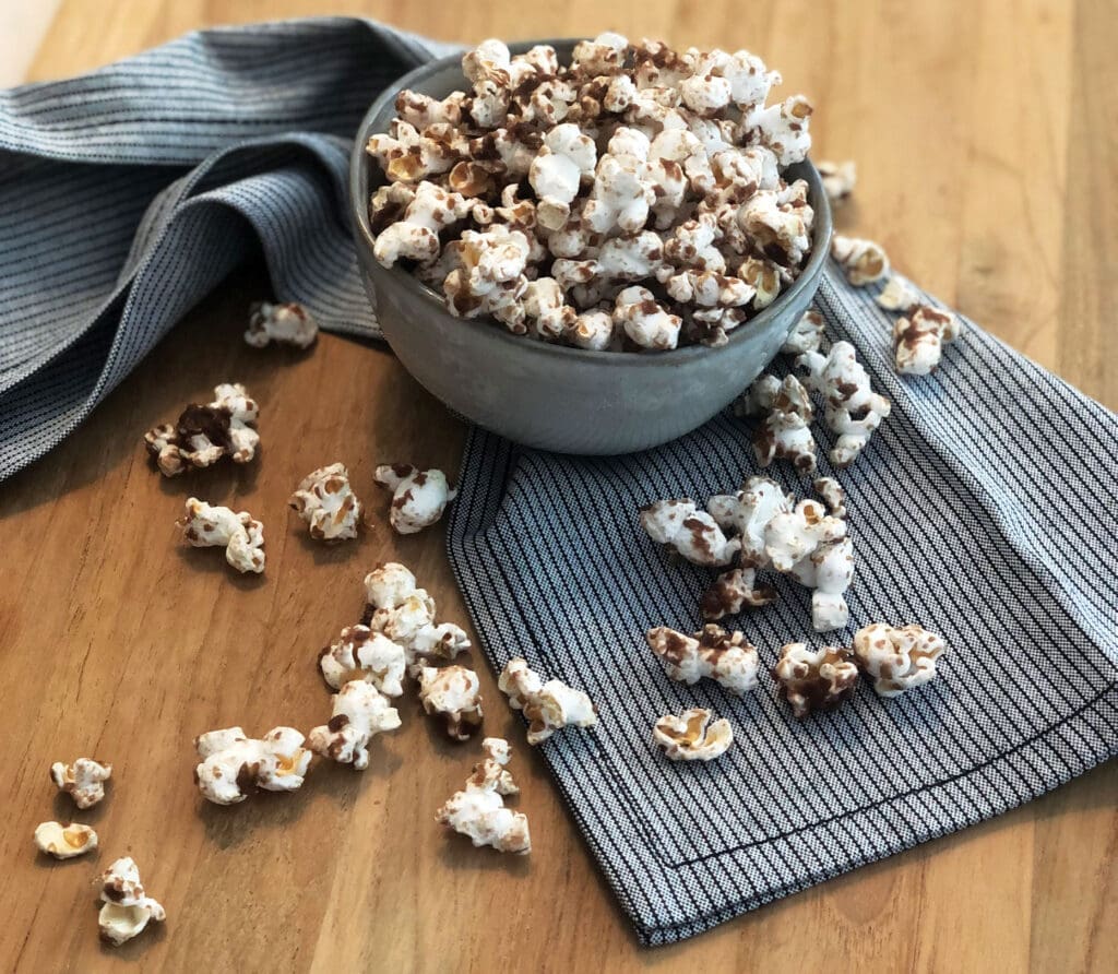A gray bowl filled with chocolate-drizzled popcorn sits on a wooden table, surrounded by scattered pieces of popcorn and jelly beans. A striped cloth napkin lies under the bowl and extends across the table.