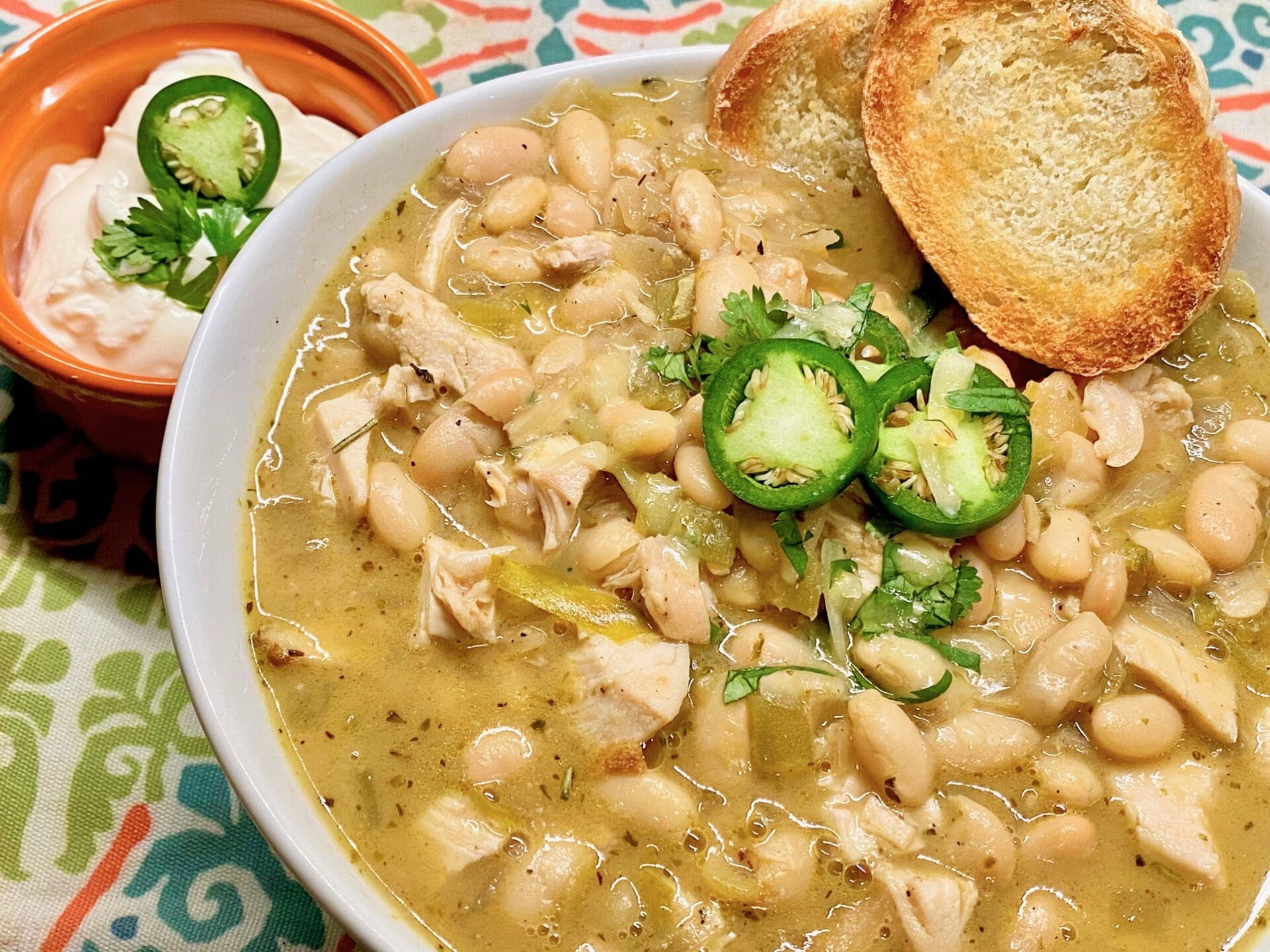 A bowl of white chicken chili featuring Latin black beans, topped with sliced jalapeños and cilantro, served alongside toasted bread. A side of sour cream garnished with cilantro sits in an orange bowl. The tablecloth boasts a colorful pattern.