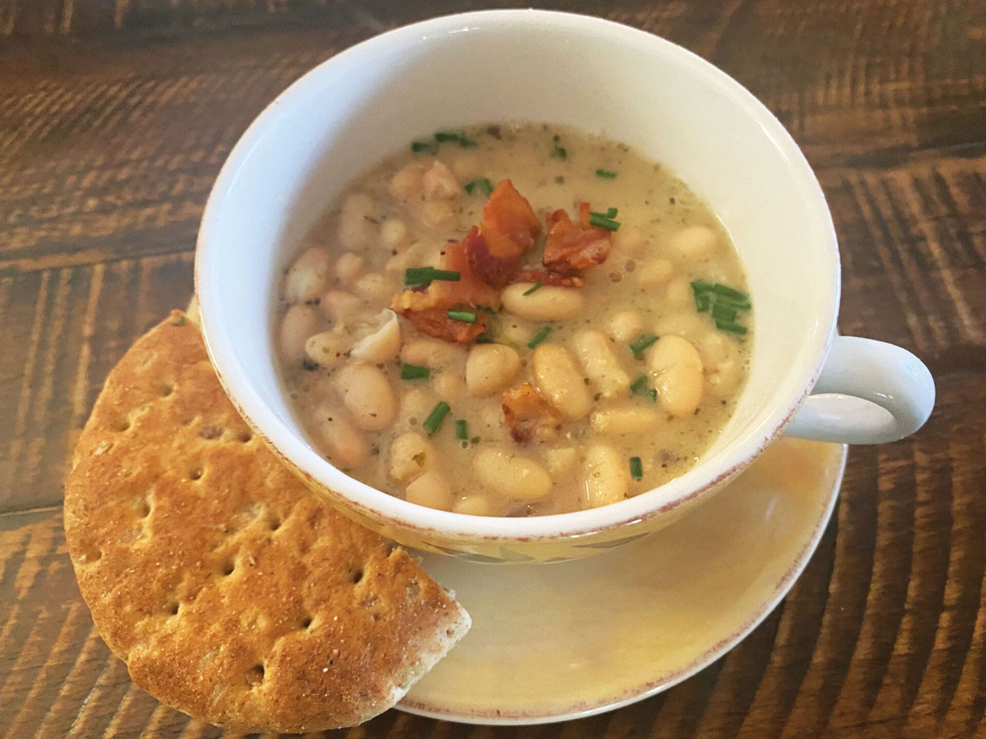 A cup of creamy white bean soup garnished with crispy bacon and chives, served alongside a saucer of cracker bread and a touch of Latin black beans. The wooden table background adds warmth to the presentation.