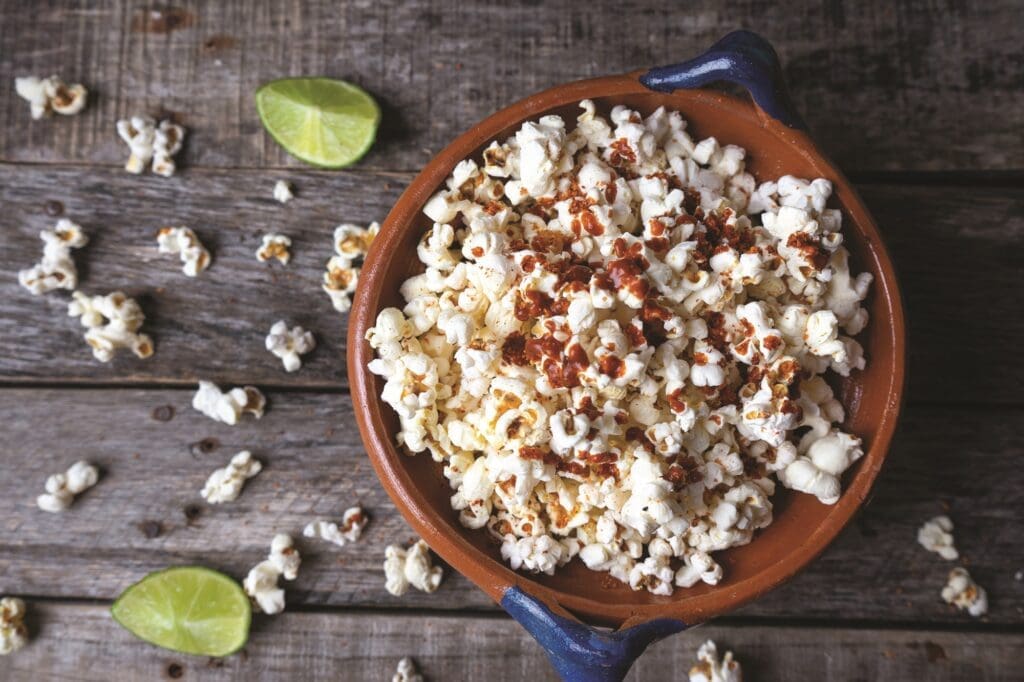 A bowl of popcorn sprinkled with chili powder sits on a rustic wooden surface, reminiscent of Latin flavors. Lime wedges and scattered popcorn pieces are around the bowl, adding a pop of color to the scene, much like a dish of black beans and rice would complement this vibrant setting.