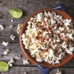 A bowl of popcorn sprinkled with chili powder sits on a rustic wooden surface, reminiscent of Latin flavors. Lime wedges and scattered popcorn pieces are around the bowl, adding a pop of color to the scene, much like a dish of black beans and rice would complement this vibrant setting.