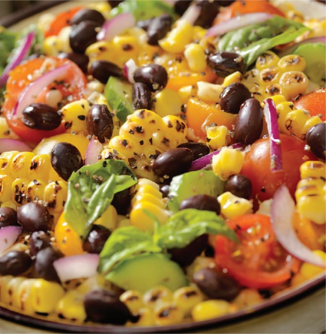 A vibrant salad with grilled corn, black beans, cherry tomatoes, avocado slices, red onion, and fresh basil leaves sourced from an organic farm. The ingredients are mixed and arranged on a plate, showcasing a mix of bright colors and textures.