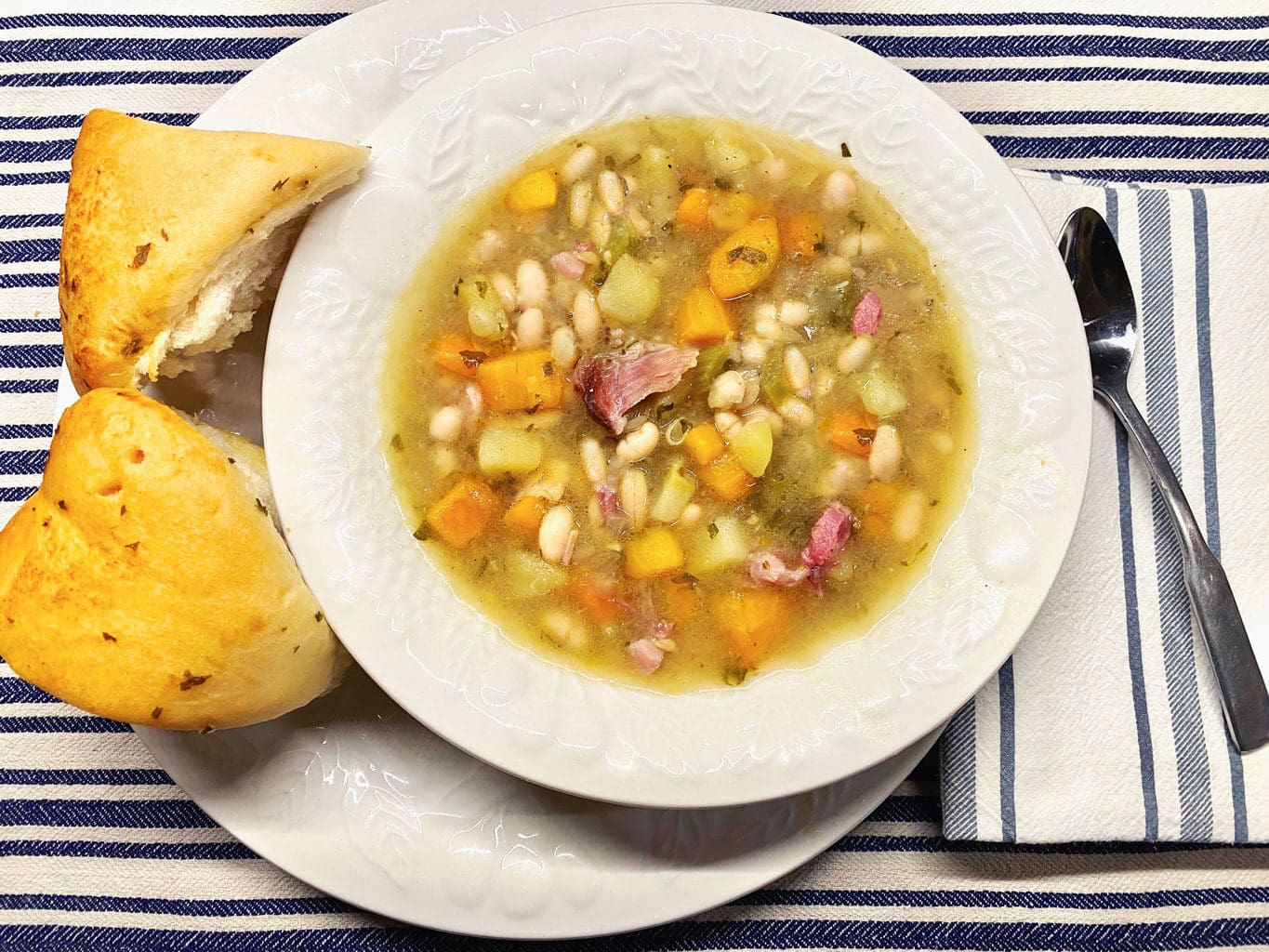 A bowl of hearty U.S. Senate bean soup with chunks of ham, carrots, and herbs sits on a white plate, accompanied by two pieces of crusty bread. A spoon and a striped napkin lie beside it on a striped blue and white tablecloth.