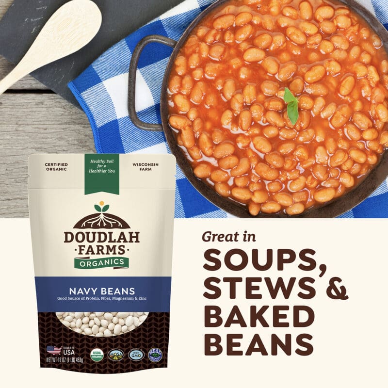 A 1LB package of Doudlah Farms Organics Navy Beans sits beside a skillet filled with cooked beans. A wooden spoon rests on a blue and white checkered cloth. Text reads: Great in soups, stews & baked beans.