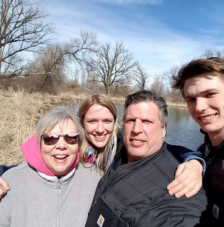 A group of four people, likely from a family farm, are smiling and taking a selfie outdoors near a small lake or pond. Leafless trees and dry grass form the backdrop under a clear blue sky. Enjoying the sunny day, they celebrate the joys of regenerative food and sustainable living.