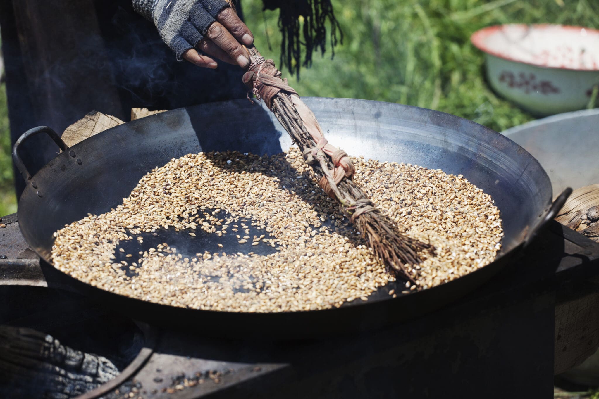 A close-up of a traditional pan roasting process for ancient grains, featuring golden grains being stirred with a handmade broom on an outdoor stove. This showcases sustainable cooking techniques and the natural preparation of nutrient-rich ancient grains for everyday recipes.