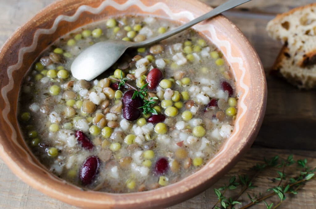 A rustic bowl of hearty soup filled with a medley of peas, red beans, and ancient grains, garnished with fresh thyme. A silver spoon rests inside the bowl, embodying everyday cooking at its finest. Slices of crusty bread are visible in the background.