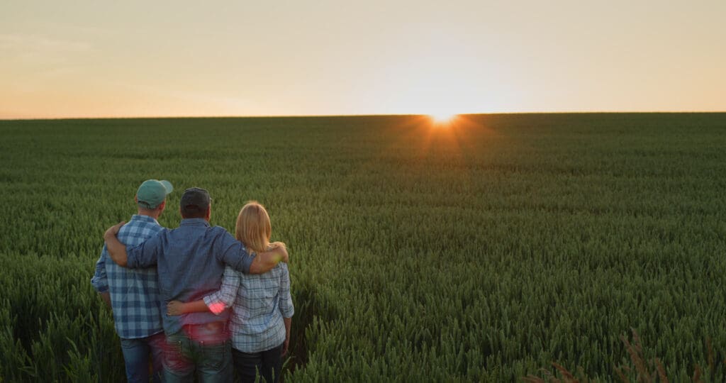 Three farmers standing arm in arm, looking at a vibrant field during sunset, symbolizing family farms' commitment to protecting the planet and sustainable agriculture.