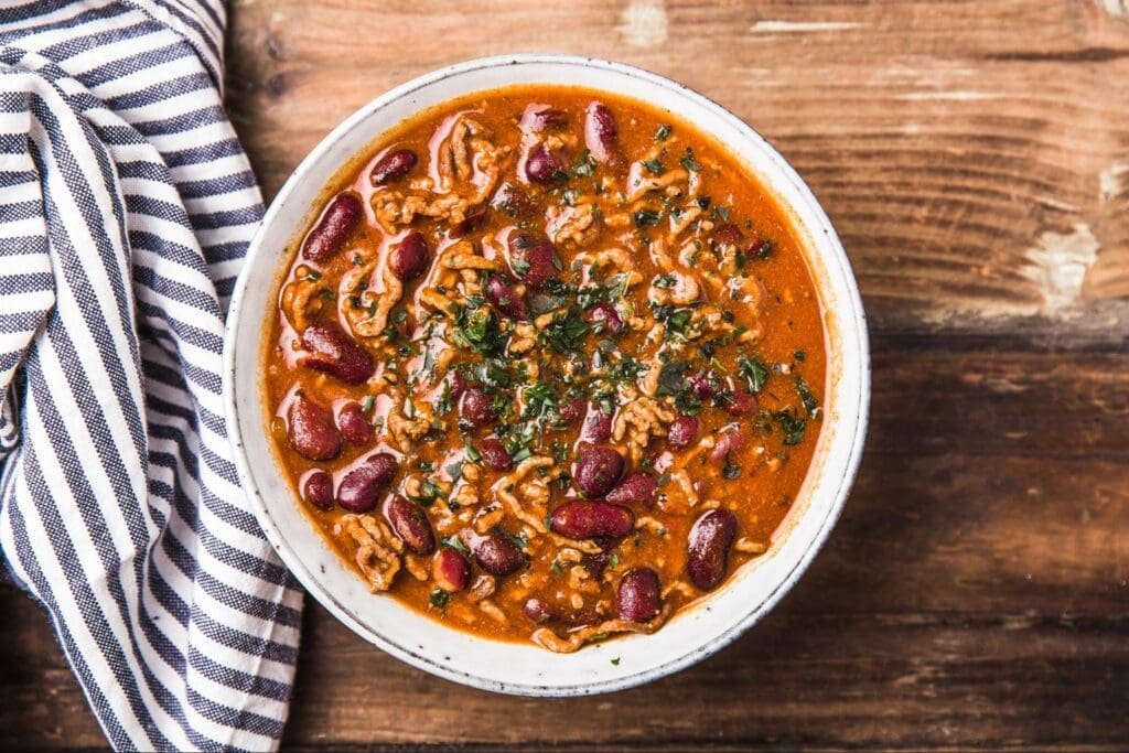 A bowl of chili with dark red kidney beans and minced meat, garnished with herbs, sits on a wooden table. A striped cloth is partially visible to the left, adding a cozy touch to this Instant Pot creation.
