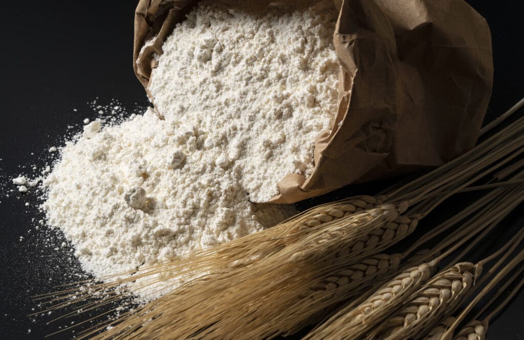 A paper bag spilling organic flour next to wheat stalks, representing the choice between organic and conventional flour.