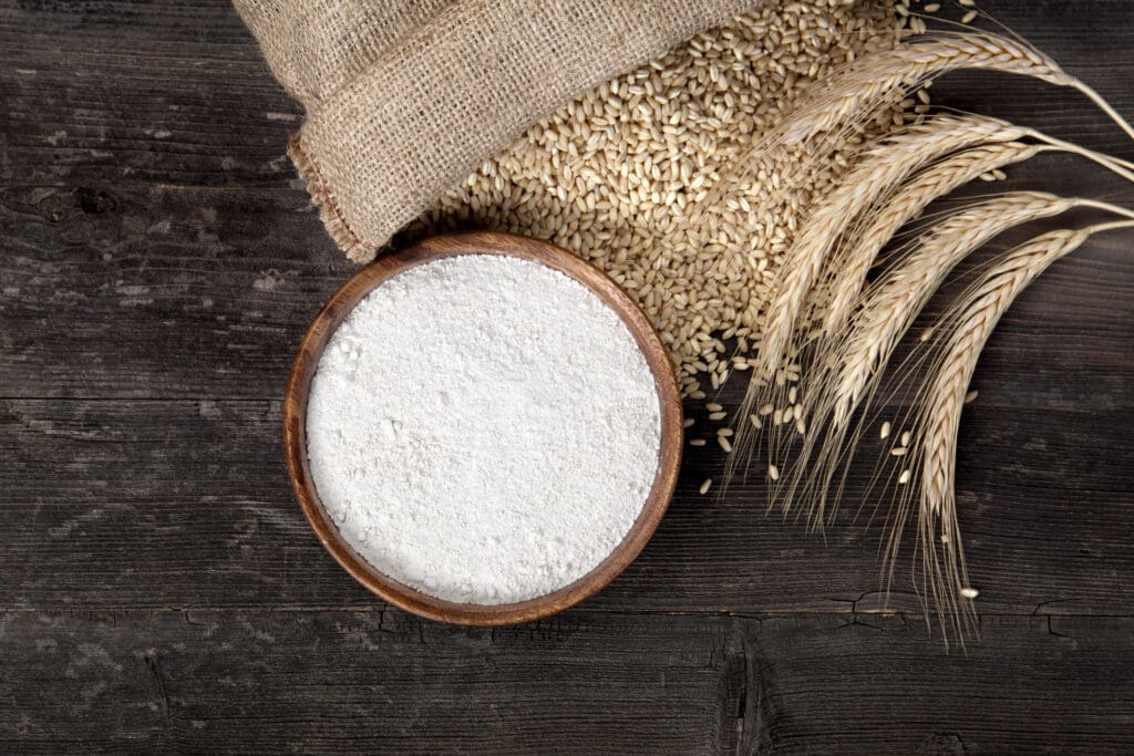 A bowl of organic flour with wheat grains and stalks on a rustic wooden surface, highlighting the benefits of organic flour over conventional flour.