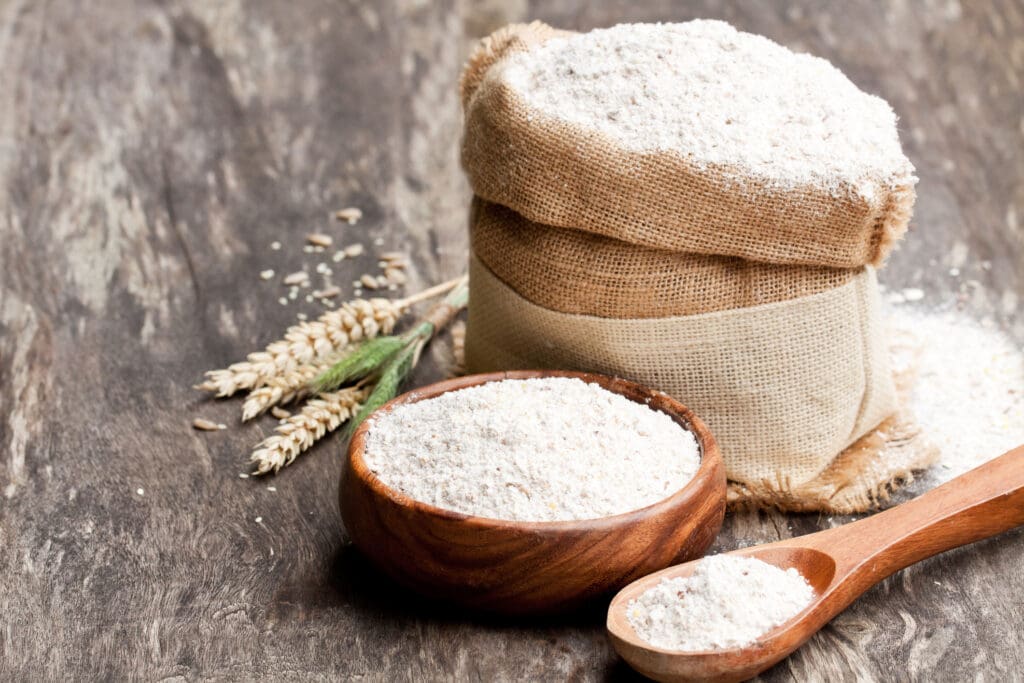 Organic flour in a wooden bowl and burlap sack, surrounded by wheat grains on a rustic wooden surface.