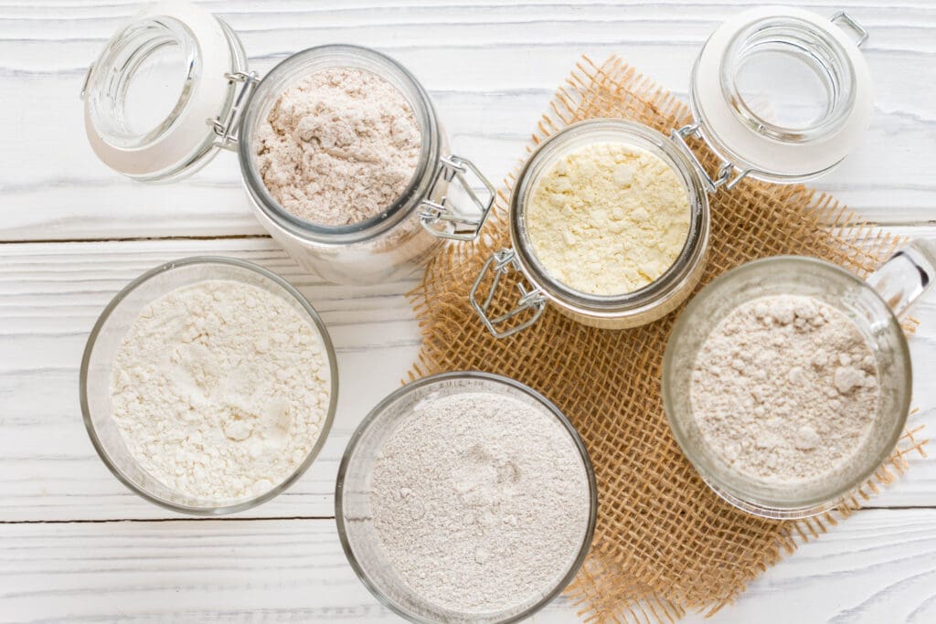 Various organic flour types in glass bowls and jars on a rustic wooden surface, highlighting unbleached and chemical-free options for healthy baking.