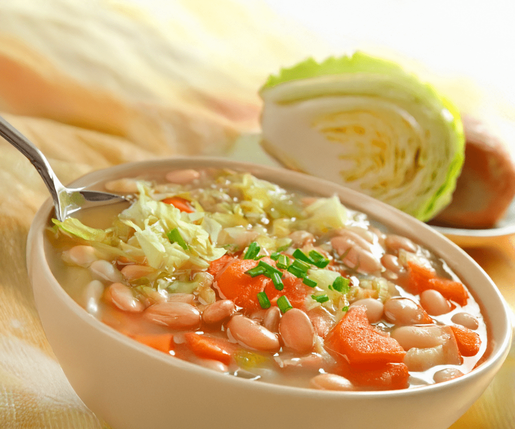 A bowl of hearty Irish white bean soup brimming with sliced carrots and chopped greens, garnished with chives. In the background, a halved cabbage rests on a light-colored tablecloth.