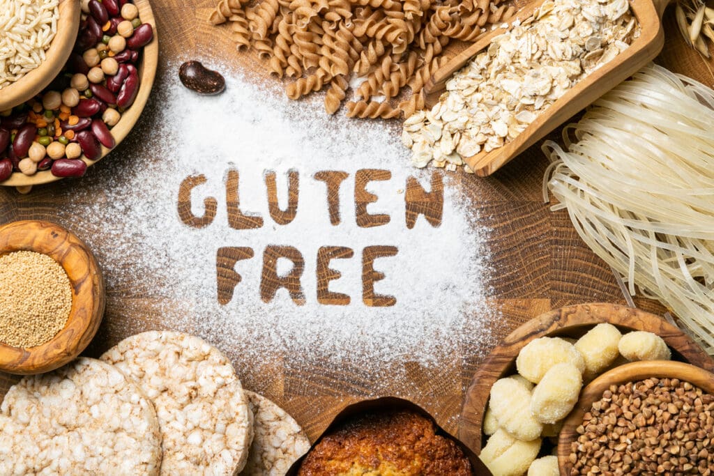 Gluten-free foods including buckwheat, rice, oats, and legumes arranged on a wooden surface with "Gluten Free" written in flour.