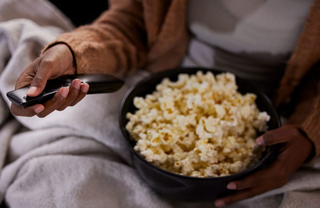 A person sits on a couch, wrapped in a blanket, holding a TV remote in one hand and a large bowl of high-fiber popcorn in the other. They appear to be ready to enjoy their fiber-rich snack while watching a show or movie.