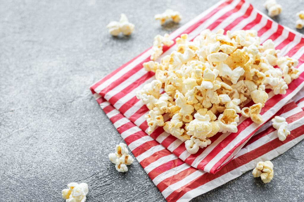 A pile of high-fiber superfood popcorn sits on red and white striped paper bags scattered across a textured gray surface, with a few fiber-rich pieces scattered around.