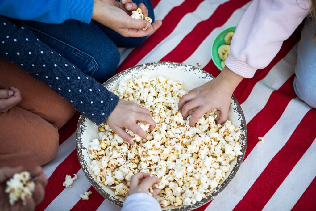 Childrens hands dive into a bowl of fiber-rich popcorn on a red and white striped blanket, with a small bowl of kiwi slices nearby.