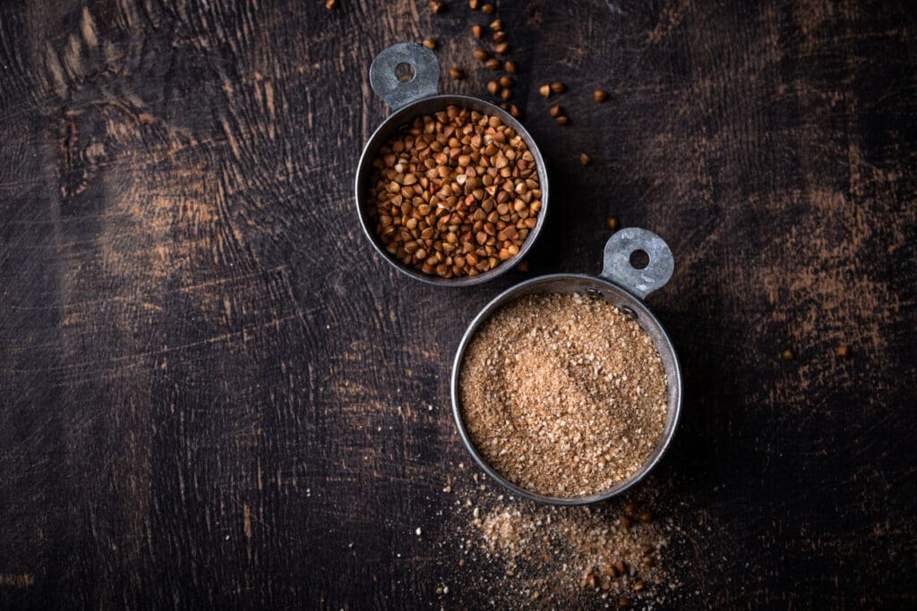 Rustic metal measuring cups filled with raw buckwheat groats and freshly milled buckwheat flour on a dark wooden surface, highlighting its gluten-free, nutrient-rich qualities.