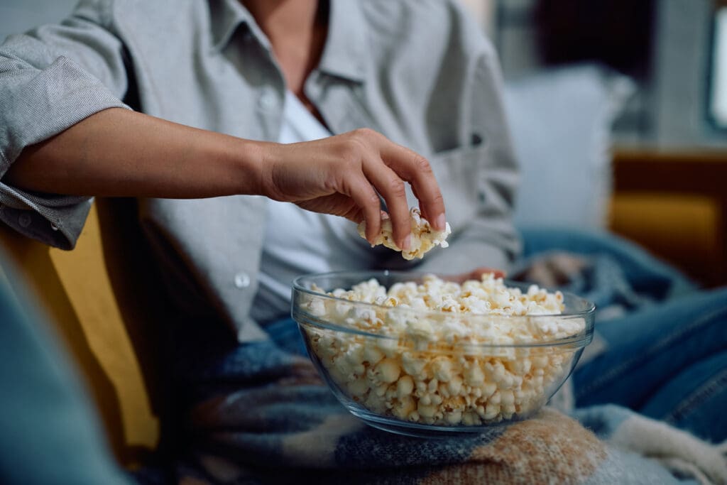 A person wearing a gray shirt is sitting on a couch, reaching for popcorn, a high-fiber superfood, from a glass bowl. A cozy blanket covers their lap, and a soft-focus background suggests a comfortable home setting.