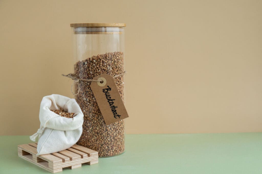 A glass jar filled with raw buckwheat groats with a rustic tag labeled "Buckwheat," alongside a small cloth sack of buckwheat on a wooden pallet, highlighting its gluten-free and nutritious qualities.