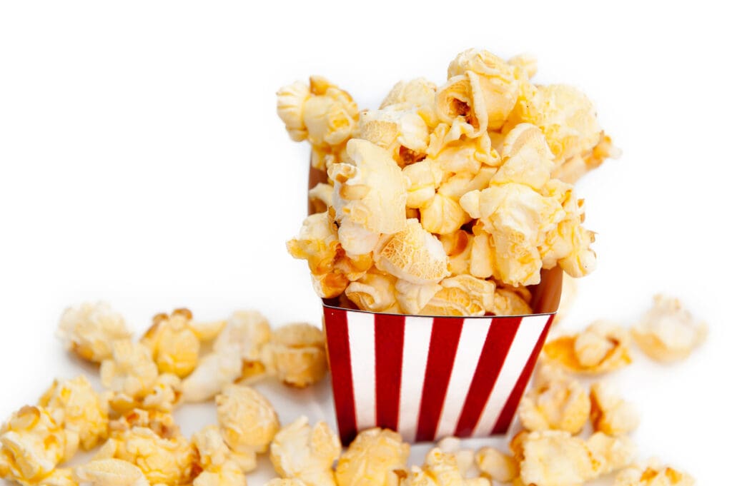 A striped red and white popcorn container brims with buttery, fiber-rich popcorn, a snack celebrated as a high-fiber superfood. Some kernels spill over playfully around the container, set against a pristine white background that emphasizes their tempting allure.