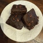 Three flour-free black bean brownies with chocolate chips are stacked on a white patterned plate, placed on a wooden table. Kid-tested and irresistibly delicious, these treats redefine indulgence without compromise.