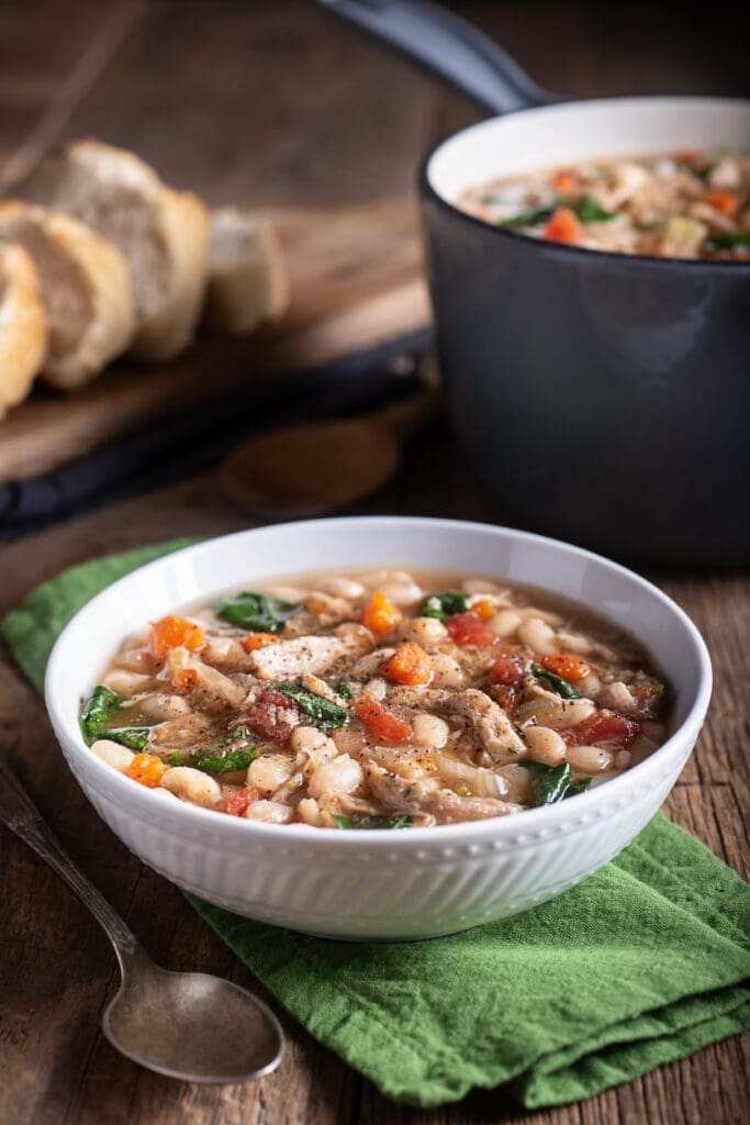 A bowl of hearty Navy Bean Soup with spinach, carrots, and chicken cozies up on a green napkin. Nearby, a spoon awaits on the wooden table. In the background, a pot simmers enticingly next to sliced bread from Doudlah Farms for perfect pairing.