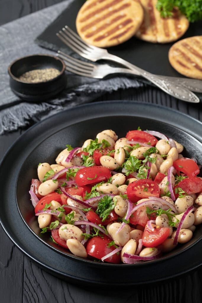Black bowl with a white bean salad featuring cherry tomatoes, red onion slices, and parsley. A small dish of pepper, two forks, and grilled pita bread on a slate tray are in the background, all set on a dark wooden table.