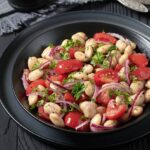 Black bowl with a white bean salad featuring cherry tomatoes, red onion slices, and parsley. A small dish of pepper, two forks, and grilled pita bread on a slate tray are in the background, all set on a dark wooden table.