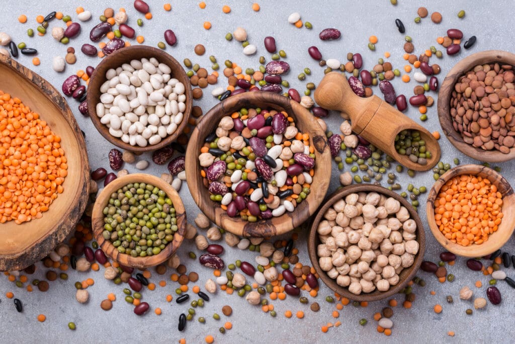 An assortment of dried beans, lentils, and legumes—great for gut health and digestion—are displayed in wooden bowls on a light surface, with chickpeas, kidney beans, white beans, and red and green lentils scattered around.