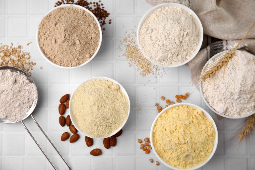 A selection of different flours in bowls on a white tiled surface caters to all your baking needs. Various grains and almonds are scattered around, with a kitchen towel and wheat stalks nearby. A metal measuring spoon with organic flour is on the left.