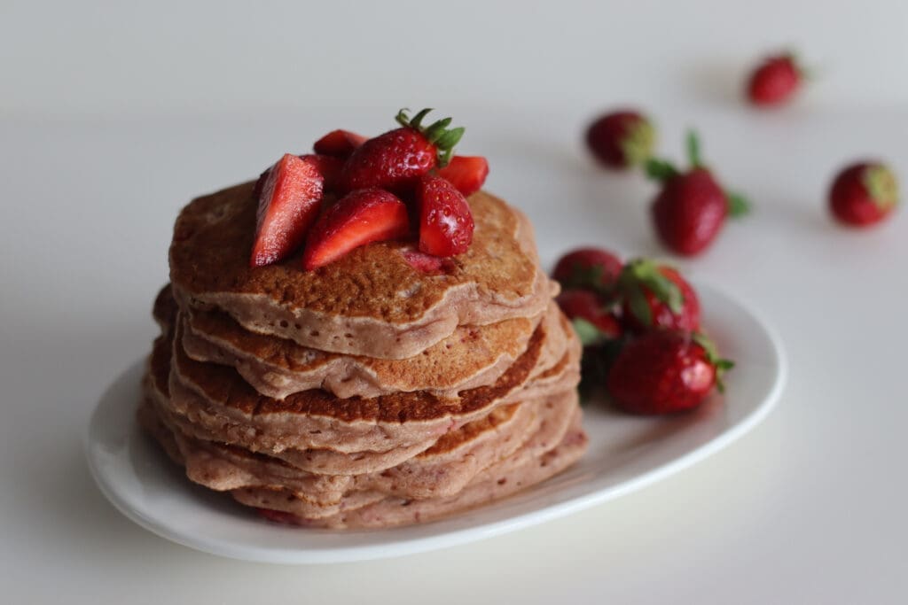 A stack of buckwheat pancakes topped with sliced strawberries sits on a white plate, while whole strawberries are artfully scattered across the white surface in the background.
